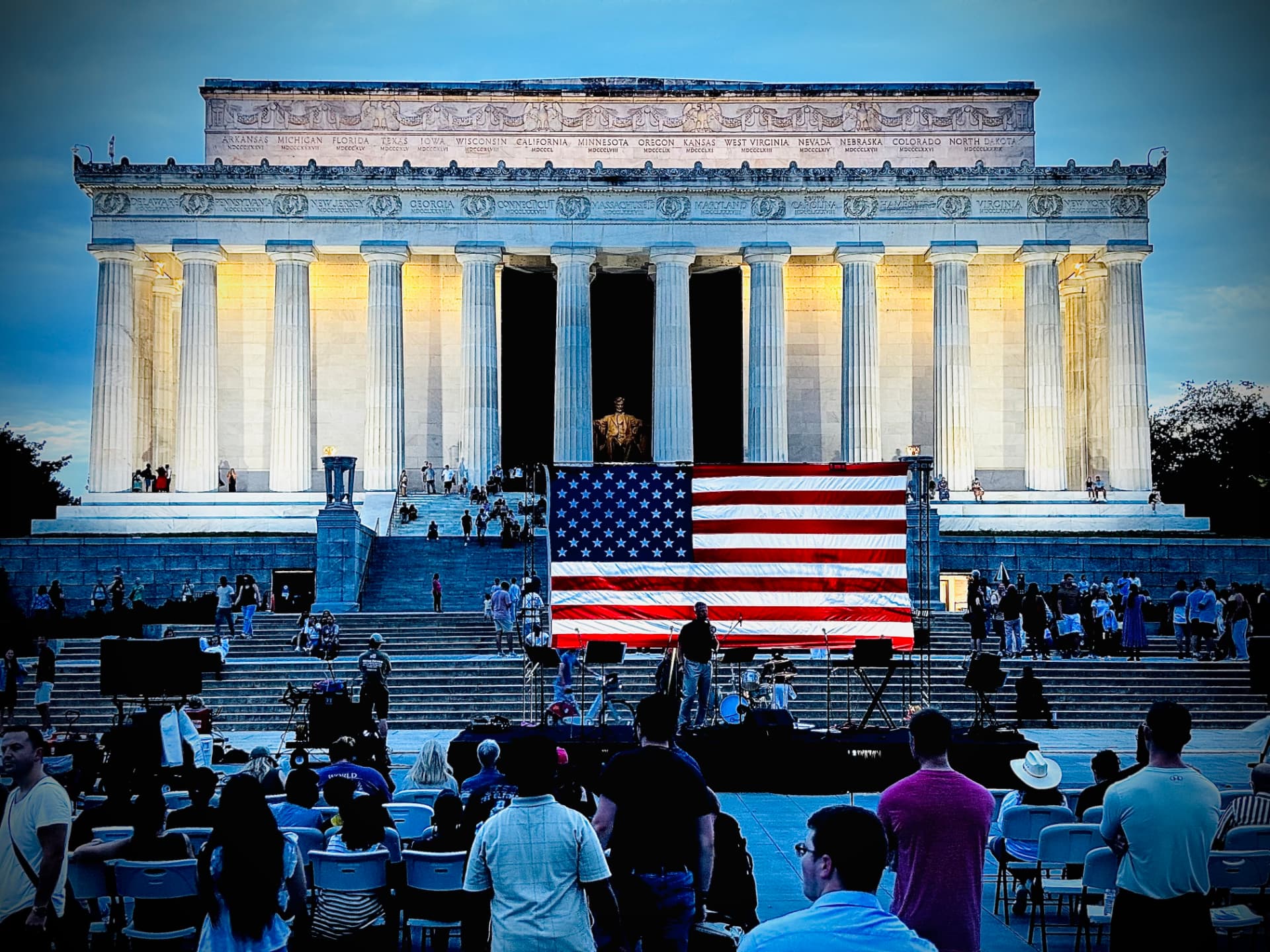 A crowd gathered before a large American flag on the steps of the Lincoln Memorial at dusk during God and Country
